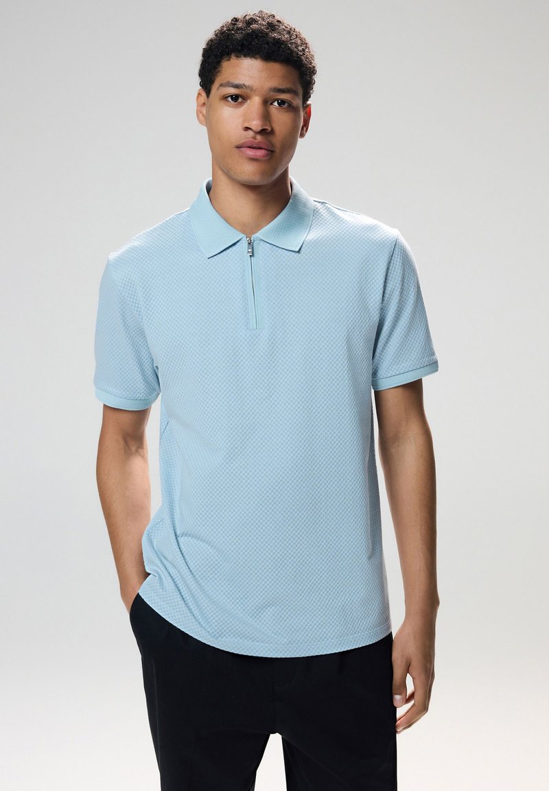 Polo shirt azzurro chiaro con colletto a zip, caratterizzato da un motivo testurizzato. Maniche corte e vestibilità comoda. Indossato con pantaloni scuri.