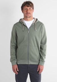 Haut à fermeture éclair vert avec une doublure gris clair, poches avant et cordon de serrage. Texture douce, coupe décontractée et logo visible sur la poitrine.