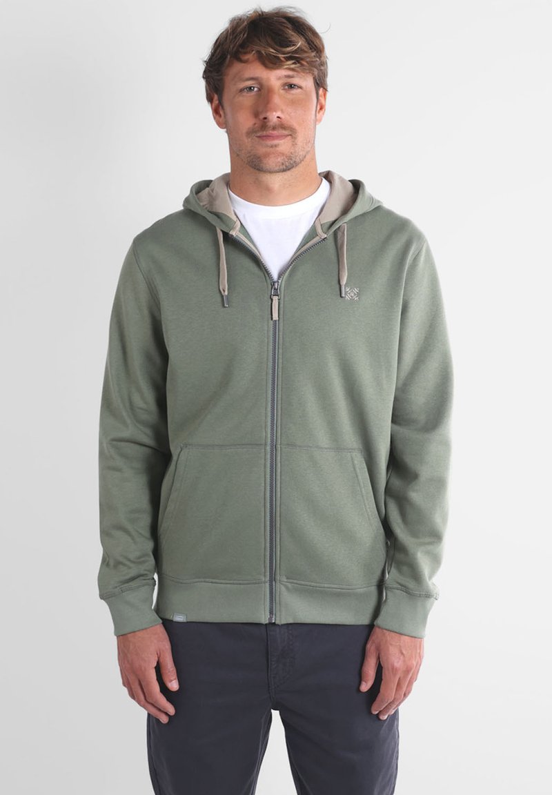 Haut à fermeture éclair vert avec une doublure gris clair, poches avant et cordon de serrage. Texture douce, coupe décontractée et logo visible sur la poitrine.
