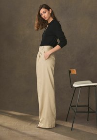 Kvinde med brunt hår iført sort langærmet top og højtaljede beige brede bukser, står ved siden af en høj stol mod en brun baggrund.