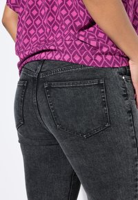Pantalones vaqueros ajustados negros con textura acanalada vertical, con dos bolsillos traseros y costuras de contraste. Combinados con una blusa morada con estampado.