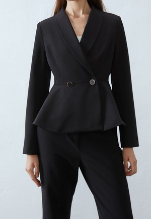 Vrouw draagt een aansluitende zwarte blazer met twee ronde knopen en bijpassende zwarte broek, staand tegen een effen lichte achtergrond.