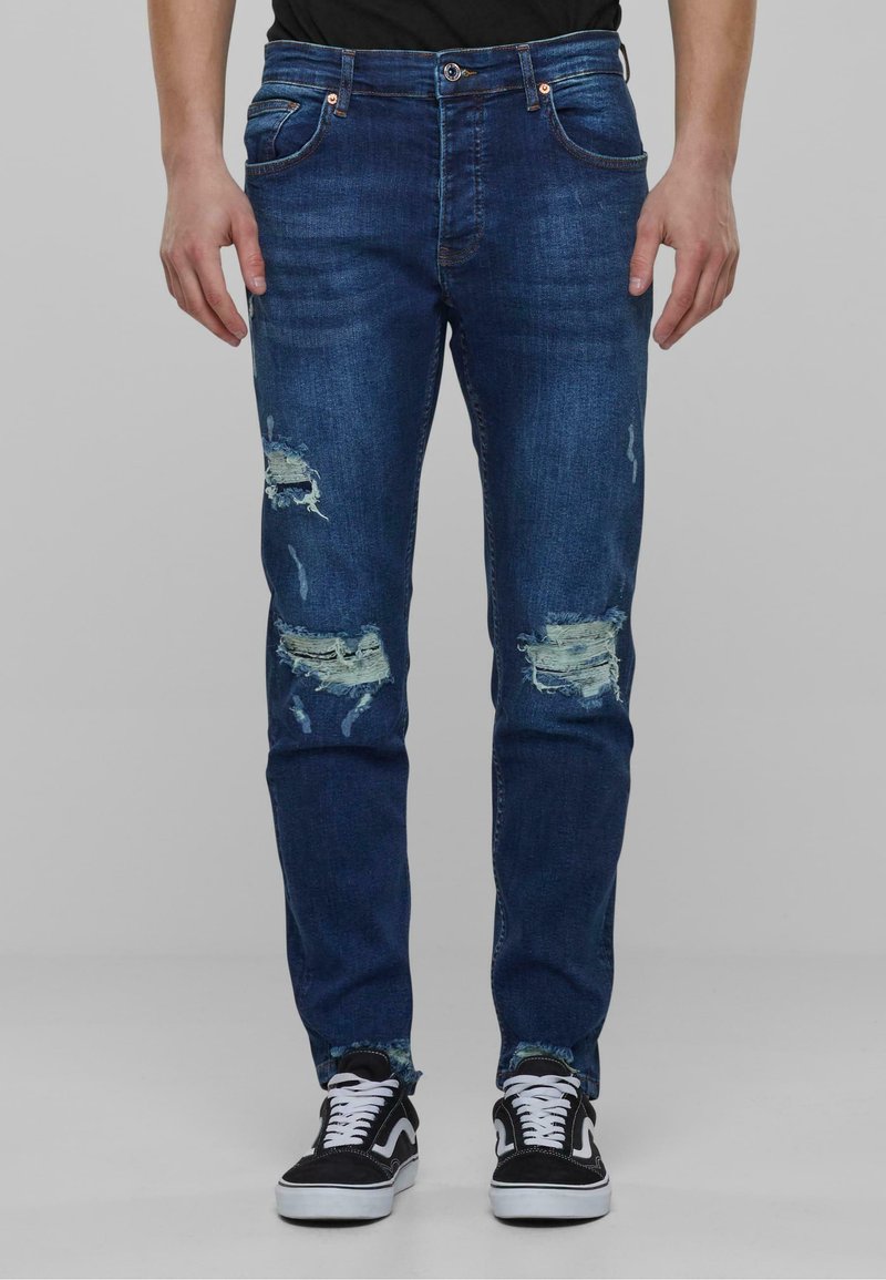 Jeans bleu foncé avec des empiècements usés, coupe droite, design à cinq poches, fermeture à bouton et coutures contrastées. Associés à des baskets noires.