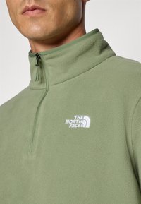 Muškarac nosi svijetlozelenu flis jaknu s četvrtastim zatvaračem i bijelim logotipom The North Face na prsima.