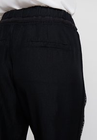 Widok z tyłu osoby noszącej czarne luźne spodnie z elastycznym pasem i rozcięciami z tyłu, w połączeniu z białą bluzką.