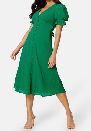Femme portant une robe midi verte avec manches bouffantes et détails de boutons, associée à des sandales à talons hauts beiges sur fond blanc.