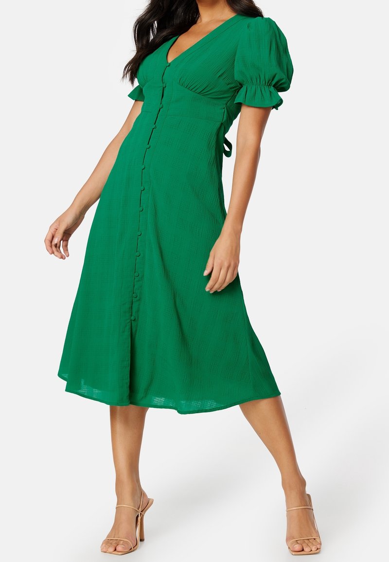 Femme portant une robe midi verte avec manches bouffantes et détails de boutons, associée à des sandales à talons hauts beiges sur fond blanc.