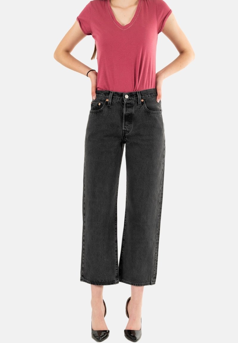 Femme portant un jean noir taille haute à jambes larges et coupé, un haut rose à manches courtes et col en V, et des talons noirs à bout pointu, mains sur les hanches.