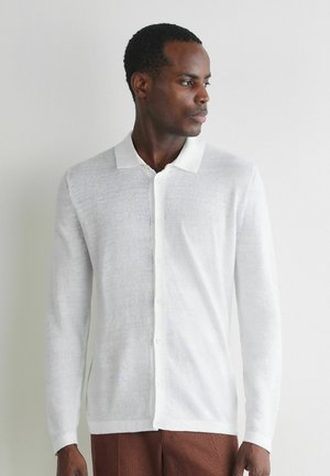 Mann trägt weißes, langärmeliges Poloshirt mit Knöpfen und braune Hose, blickt nach rechts vor einem einfarbig hellen Hintergrund.