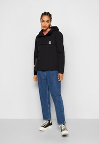 Giacca nera con cappuccio e tasca frontale, abbinata a jeans blu e sneaker alte nere. Presenta un patch con logo sul petto.