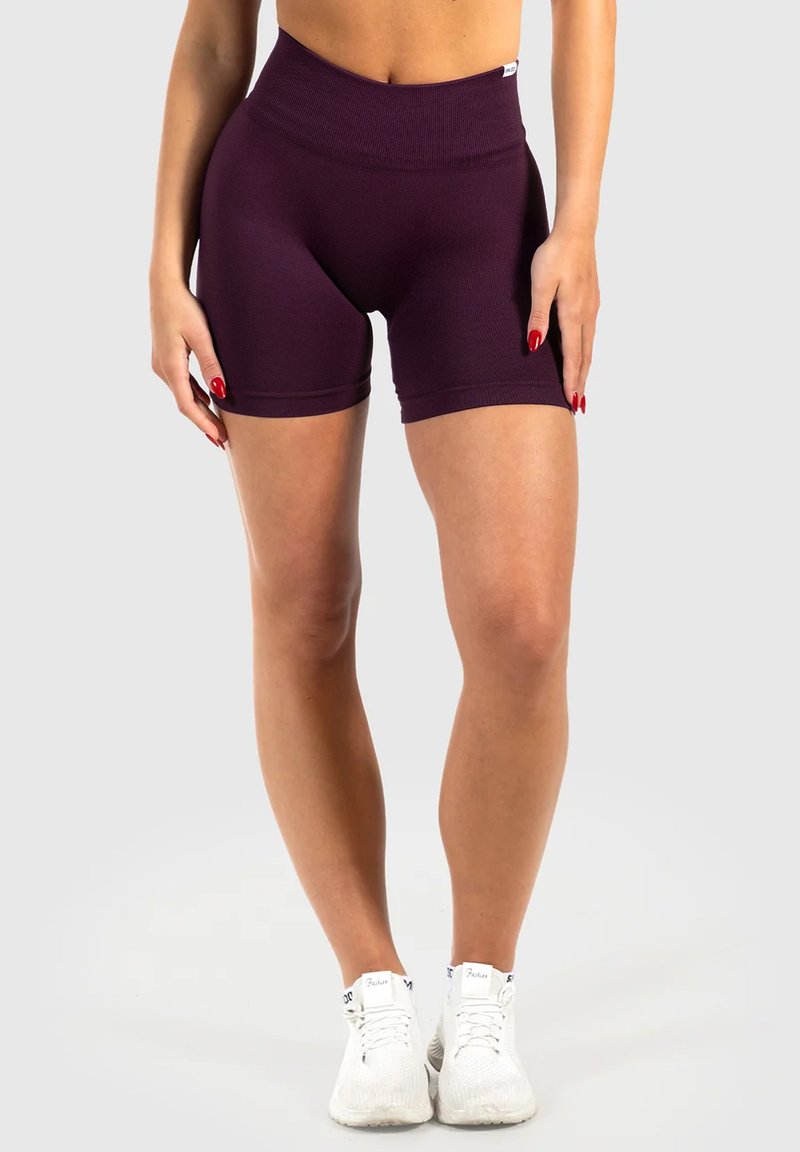 Femme portant un short de cycliste taille haute violet foncé et des baskets blanches, debout devant un fond uni.