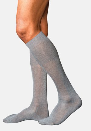 Beine mit hellgrauen knielangen Kompressionssocken, stehend mit einem Fuß flach und dem anderen leicht auf den Zehen erhoben.