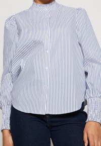 Blouse boutonnée à rayures verticales blanches et bleues, avec un col montant et des poignets smockés. Fabriquée en tissu léger.