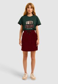 Maglietta verde con il testo "SWEET ESCAPE", abbinata a una gonna in velluto a coste bordeaux con vita regolabile e tasche frontali. Sneakers bianche.