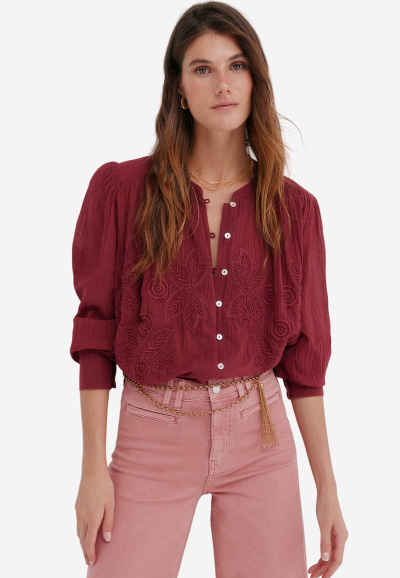 Blouse bordeaux à texture avec des manches bouffantes, présentant une broderie florale et une fermeture à boutons ; associée à un pantalon ajusté rose clair.