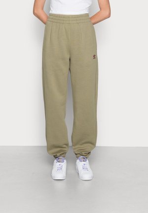 Person iført beige Adidas joggingbukser med elastiske manchetter og hvide Adidas sneakers, stående mod en ensartet lys baggrund.