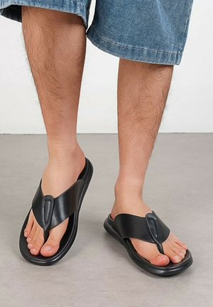 Pieds masculins portant des sandales en cuir noires de type tongs assorties à un short en denim bleu sur un sol gris clair avec un fond blanc.