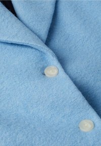 Gros plan sur un tissu bleu clair à la texture douce avec deux boutons ronds translucides et un détail de col plié.