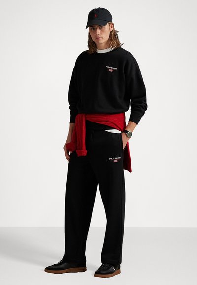 Tânăr îmbrăcat cu un hanorac și pantaloni Polo Sport negri, cu o șapcă neagră, un pulover roșu legat în jurul taliei, stând cu o mână în buzunar.