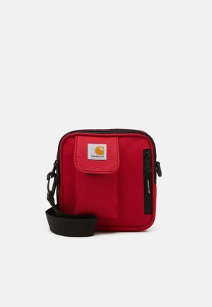 Petit sac bandoulière rouge en tissu avec poche à rabat avant, poche zippée verticale, sangle noire réglable et écusson logo Carhartt.
