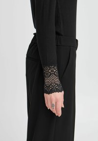 Schwarzes Langarmoberteil mit geripptem Textur und Spitzenbesätzen an den Ärmeln. Der Ärmelaufschlag weist filigrane Blumenmuster und gewellte Kanten auf.