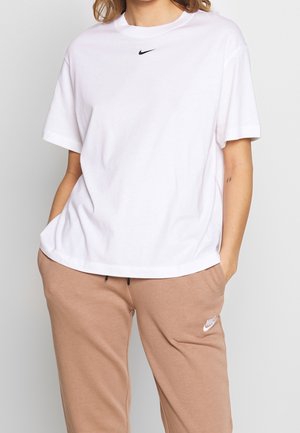 Person trägt ein weißes Nike-T-Shirt und beige Nike-Sweatpants mit den Händen in den Taschen vor einem einfarbigen hellen Hintergrund.