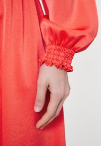Helles rotes Kleidungsgewebe mit glatter Textur, das ein gerafftes elastisches Manschettendetail am Ärmel und ein gerafftes Oberteil aufweist.