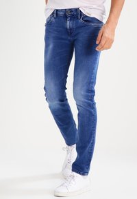 Úzké modré džíny s vybledlým povrchem, které mají pět kapes a přední zapínání na knoflík. V kombinaci s bílými teniskami.