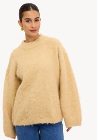 Pull en peluche beige avec un col haut et des manches longues, présentant une finition texturée. Porté sur un jean en denim bleu.
