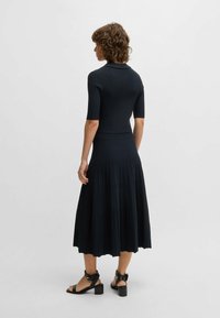 Czarna sukienka z prążkowanej tkaniny z dopasowaną górą i plisowanym dołem, trzema czwartymi rękawami oraz kołnierzykiem. Połączona z czarnymi sandałami na słupku.