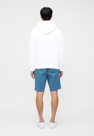 Άνδρας με λευκό φούτερ με κουκούλα, μπλε σορτς και λευκά παπούτσια slip-on, στέκεται με την πλάτη προς το μέρος μας μπροστά σε ένα απλό φωτεινό φόντο.