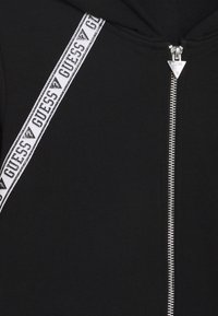Sudadera negra con cremallera y una tira blanca con el logo repetido de "GUESS" a lo largo del hombro. Fabricada en suave algodón.