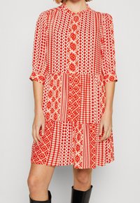 Robe à motifs rouges avec des manches trois-quarts, fermeture à boutons sur le devant et une jupe évasée. Présente des designs géométriques et floraux dans différentes nuances de rouge.