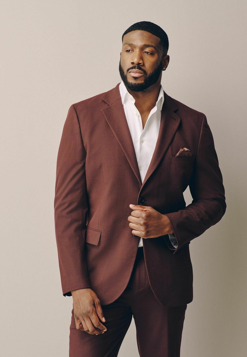 Homme barbu portant une veste de costume marron et une chemise blanche, ajustant sa veste tout en regardant sur le côté sur un fond uni.