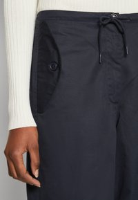 Un pantalon bleu marine avec une texture lisse, un cordon de serrage réglable et des poches latérales avec des rabats arrondis et des fermetures à bouton.