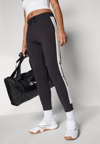 Svarte joggebukser med hvite sidestriper, elastisk midje og ribbede ankler. Kombinert med en svart treningsbag og hvite sportssko.