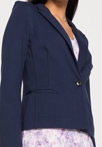 Žena v přiléhavém námořnickém blejzru přes levandulové květinové šaty s výstřihem do V, zobrazená od hrudníku po pás.