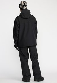 Veste extérieure noire imperméable avec capuche, assortie à un pantalon noir. Présente un design minimaliste, des manches longues et un tissu texturé.