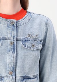 Ανοιχτό μπλε τζιν μπουφάν με ασημί κούμπωμα, μπροστινές τσέπες και κεντημένο λογότυπο. Φορεμένο πάνω από ένα κόκκινο μπλουζάκι. Υφασμάτινη επιφάνεια με φθαρμένα σημεία.