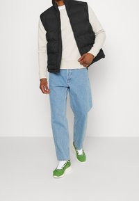 Czarna pikowana kamizelka na jasnoszarej bluzie, zestawiona z jasnoniebieskimi dżinsami i zielonymi trampkami z białymi sznurowadłami oraz teksturowanymi materiałami.