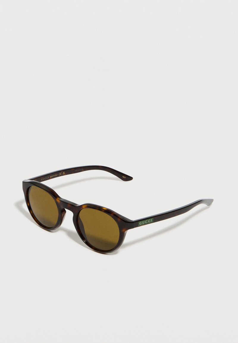 Lunettes de soleil rondes en écaille de tortue avec verres bruns et logo Gucci vert sur des branches fines, présentées sur une surface blanche.