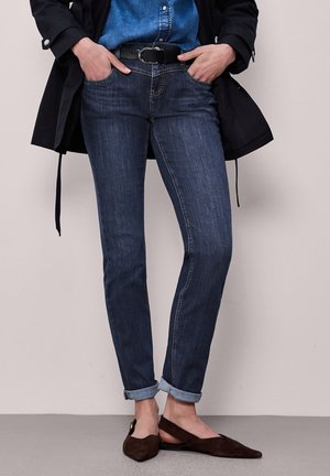 Jeans slim fit blu scuro risvoltati alle caviglie, abbinati a una cintura nera, camicia di denim, giacca nera e ballerine marroni con punta appuntita.