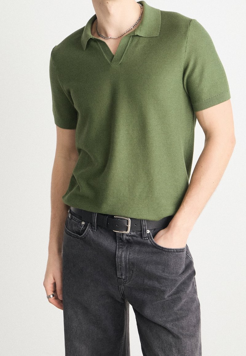 Homme portant un polo en maille vert olive à manches courtes, ceinture en cuir noir et jean gris foncé, avec une main dans la poche et un collier argenté.