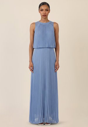 Woman wearing sleeveless light blue pleated maxi dress standing against a plain beige background.