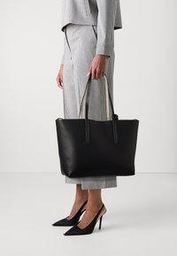 Czarna skórzana torba na ramię o fakturze z drobnych wypukłości, z długimi uchwytami i kontrastowym jasnym wnętrzem. Charakteryzuje się minimalistycznym designem.