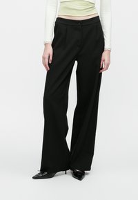 Kvinne iført høy midje, vide svarte bukser kombinert med svarte spisse hæler og en kremfarget langermet topp, stående mot hvit bakgrunn.