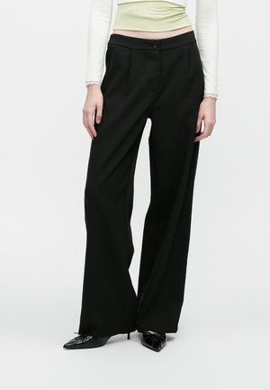 Femme portant un pantalon noir taille haute à jambes larges associé à des talons noirs à bout pointu et un haut à manches longues crème, se tenant devant un fond blanc.