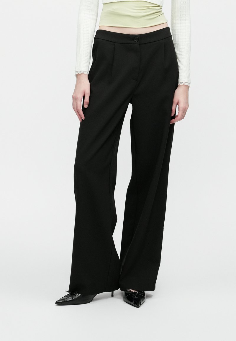 Kvinne iført høy midje, vide svarte bukser kombinert med svarte spisse hæler og en kremfarget langermet topp, stående mot hvit bakgrunn.