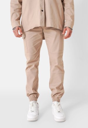 Beige cargobroek met elastische boorden, meerdere zakken en een gladde, lichte stof; gecombineerd met witte sneakers.