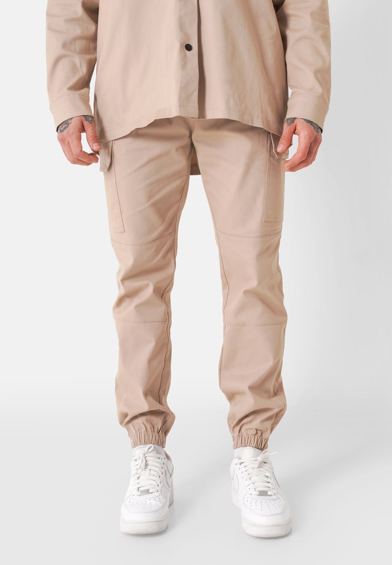 Beige cargobroek met elastische boorden, meerdere zakken en een gladde, lichte stof; gecombineerd met witte sneakers.